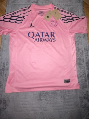 PSG x Jordan rosa matchtröja - Säljer en rosa PSG x Jordan matchtröja med svarta detaljer och vingmönster på axlarna. Tröjan har tryckt Qatar Airways-logga på bröstet och Paris Saint-Germain-emblem. Perfekt för fotboll eller streetwear.