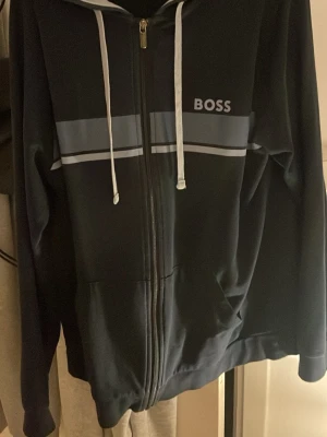 Marinblå hoodie från BOSS inkluvise byxor till - Snygg marinblå hoodie från BOSS med dragkedja framtill, vita dragsnören och två breda blå ränder över bröstet. Stort BOSS-tryck på vänster sida. Hoodien har fickor på sidorna och är tillverkad i mjukt material.