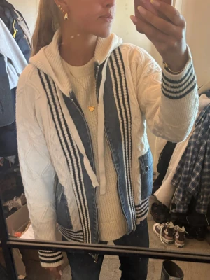 Vit stickad hoddie jeansjacka  - Säljer en vit stickad hoodie med blå jeansdetaljer och dragkedja framtill. Tröjan har ribbade muddar med blå ränder, fickor och huva med snören. Snygg kabelstickning på ärmarna och en avslappnad passform. Finns ingen storlek men passar från S-L 