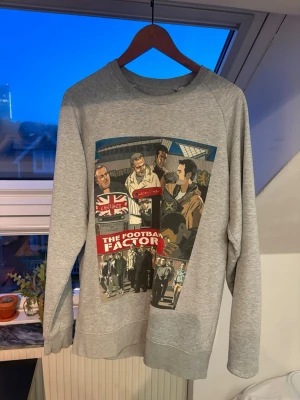 Grå sweatshirt The Football Factory - Säljer en grå sweatshirt med stort färgglatt tryck på framsidan från filmen The Football Factory. Tröjan har rund halsringning och ribbade muddar vid ärmslut och nederkant. Perfekt för dig som gillar streetwear och filmreferenser.