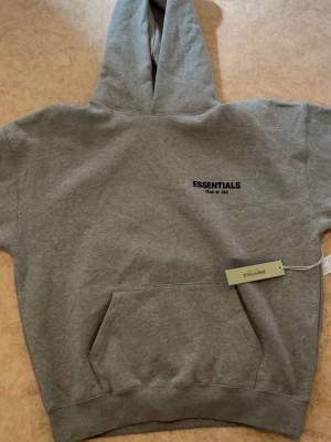 Grå Essentials hoodie  - Snygg grå hoodie från Fear of God Essentials med korta ärmar och stor huva. Tryck med 'ESSENTIALS FEAR OF GOD' på både bröstet och ryggen. Klassisk känguruficka framtill och ribbade muddar. Perfekt för en avslappnad streetwear-look.