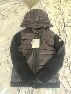 Svart pufferjacka från Moncler - Snygg svart pufferjacka från Moncler med quiltad front och huva. Ärmarna är i stickat material och jackan har dragkedja framtill. Klassisk Moncler-logga på ärmen och praktiska fickor..