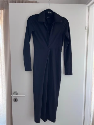 Svart långärmad klänning - Snygg svart klänning från Gina med lång ärm och markerad krage. Modellen har en djup v-ringning och är tillverkad i ett ribbat, stretchigt material som sitter skönt på kroppen. Perfekt för dig som gillar stilrena och trendiga plagg.