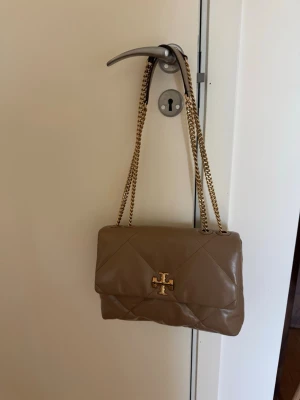 Beige axelremsväska från Tory Burch - Snygg beige handväska från Tory Burch i mjukt quiltat läder med guldfärgad kedjerem och logga framtill. Väskan har ett stort fack, innerficka med dragkedja och magnetlås. Perfekt för dig som gillar stilrena accessoarer med lyxig känsla.