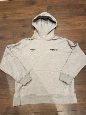 Grå hoodie från Levi's med tryck - Snygg ljusgrå hoodie från Levi's med svart tryck på bröstet och texten 'The Original' på ena sidan. Klassisk känguruficka framtill och ribbade muddar. Perfekt för en avslappnad streetstyle-look.