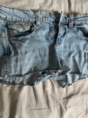 Blå jeansshorts med fransig kant - Snygga blå jeansshorts med rå, fransig kant och klassisk femficksdesign. Shortsen har låg midja och är perfekta för varma dagar. De är tillverkade i bomull och har en avslappnad passform.