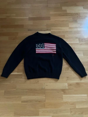 Mörkblå stickad tröja Ralph Lauren - Snygg mörkblå stickad tröja från Ralph Lauren med amerikansk flagga på bröstet och RL-broderi. Tröjan har rund halsringning och ribbstickade muddar vid ärmslut och nederkant. Perfekt för dig som gillar klassisk och preppy stil. 