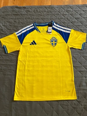 Svensk landslagströja Adidas gul/blå - Gul och blå fotbollströja från Adidas med Sveriges landslagsemblem på bröstet. Tröjan har korta ärmar, blå detaljer på axlar och ärmslut samt klassiska Adidas-ränder. Tillverkad i lätt och ventilerande material.