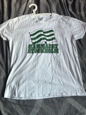 Vit Hammarby t-shirt med tryck - Vit t-shirt med grönt Hammarby Stockholm-tryck och två gröna flaggor på bröstet. Klassisk passform och rund halsringning. Perfekt för dig som vill visa ditt stöd för Hammarby. Materialet känns som bomull och t-shirten har korta ärmar.