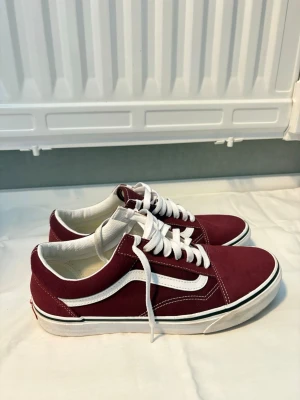 Vinröda Vans Old Skool sneakers - Vinröda Vans Old Skool sneakers med vit sula, vita skosnören och den klassiska vita sidoremsan. Ovandelen är i mocka och canvas, med låg profil och snygga kontrastsömmar. Perfekta för en streetwear-look.