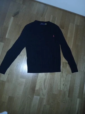 Svart stickad tröja från Ralph Lauren - Klassisk svart stickad tröja från Ralph Lauren med rund halsringning och diskret broderad logga i rött på bröstet. Tröjan har lång ärm och ribbade muddar. Perfekt för en stilren och enkel look.