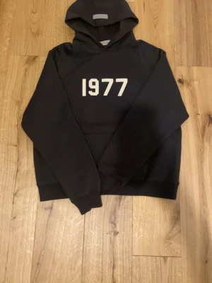 Svart hoodie från essentials 1977 - Svart hoodie med vit text '1977' på bröstet. Klassisk modell med huva och ribbade muddar. Perfekt för en avslappnad och stilren look. Materialet känns mjukt och bekvämt, passar till jeans eller joggers.