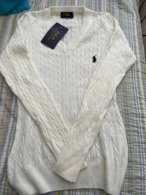 Vit kabelstickad tröja Polo Ralph Lauren - Vit kabelstickad tröja från Polo Ralph Lauren med V-ringning och klassisk broderad logga på bröstet. Långärmad modell i mjukt material, perfekt för lager-på-lager och stilrena outfits.