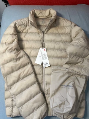 Beige pufferjacka från Uniqlo XS - Snygg beige pufferjacka från Uniqlo med PUFFERTECH-teknologi. Jackan är lätt, har hög krage och dragkedja framtill. Tillverkad i slitstarkt material och kommer med en praktisk förvaringspåse. Perfekt för kalla vinterdagar.