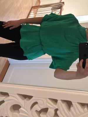 Grön volangblus med kort ärm - Snygg grön blus med volangdetalj i midjan och lös passform. Blusen har korta ärmar och är tillverkad i ett lätt, flowy tyg som ger en luftig känsla. Perfekt att styla med jeans eller kjol för en trendig look.