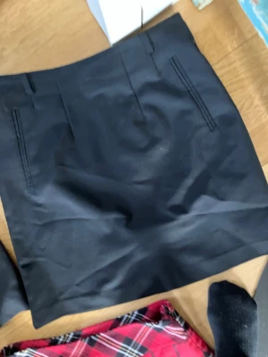 Svart kortkjol med fickdetaljer - En stilren svart kortkjol med raka linjer och diskreta fickdetaljer framtill. Kjolen har bälteshällor och är perfekt för en trendig och enkel look. Passar till många olika outfits och är lätt att matcha.