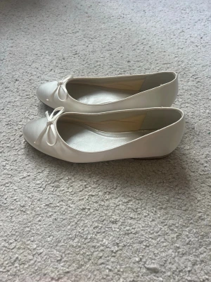 Vita ballerinaskor med rosett - Snygga vita ballerinaskor med rund tå och söt rosettdetalj framtill. Skorna har en klassisk och stilren design med låg sula och är tillverkade i ett glansigt material som ger en elegant känsla.