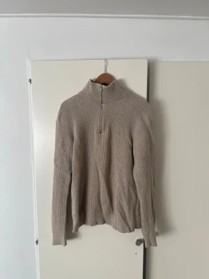 Beige ribbstickad halvzip tröja - Säljer en beige ribbstickad tröja med halv dragkedja och hög krage. Tröjan har lång ärm och avslappnad passform, perfekt för lager på lager. Materialet ser ut att vara mjukt och stretchigt, troligen bomull eller bomullsblandning. Hör av er vid frågor!