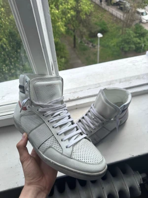 Saint Laurent vita high top sneakers - Vita high top sneakers från Saint Laurent i premiumläder med perforerade paneler och vita skosnören. Skorna har en mörkblå detalj vid hälen och en röd accentlinje. Klassisk siluett med högt skaft och diskret branding på plösen.