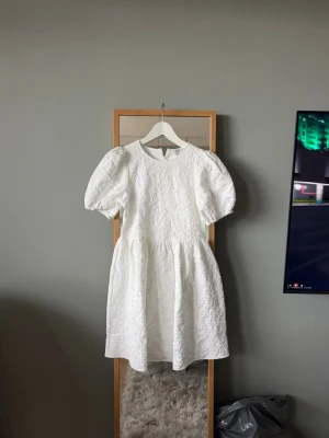 Vit kort klänning från ByIC - Superfin vit kort klänning från ByIC med puffärmar och vackert blommigt strukturmönster. Klänningen har rund halsringning och dragkedja i ryggen. Perfekt för studenten. Prislapp kvar.
