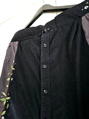 Svart manchesterkjol med broderi - Unik svart manchesterkjol med knappar framtill och gråa paneler på sidorna. Kjolen har broderade blommor och blad längs sidorna som ger en cool och kreativ vibe. Perfekt för dig som gillar detaljer och vill sticka ut.