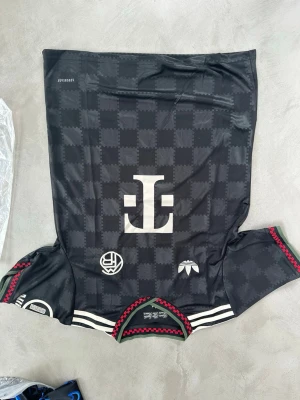 Svart Adidas x byern  matchtröja - Snygg svart matchtröja från Adidas i samarbete med byern . Tröjan har rutmönster i tyget, vita loggor och detaljer samt röda och gröna sömmar vid ärmslut och hals. Tillverkad i lätt och ventilerande polyester, perfekt för träning eller match.