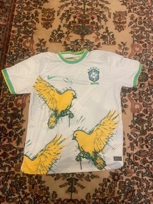Vit Brasilien fotbollströja  - Vit fotbollströja från Nike med Brasilien-emblem på bröstet och gröna detaljer vid krage och ärmslut. Framsidan har stora gula fåglar som motiv. Tröjan är kortärmad och tillverkad i ett lätt, ventilerande material.
