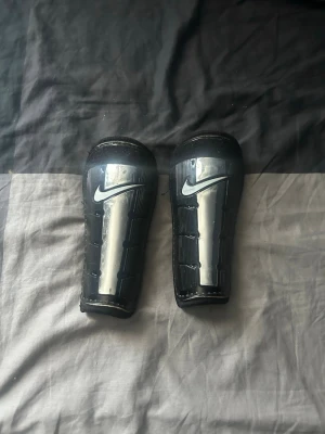 Nike benskydd svarta och silver - Snygga svarta benskydd från Nike med silverdetaljer och klassisk logga. Lätta och smidiga med hård plast på framsidan och mjuk insida för extra komfort. Perfekta för fotbollsträning eller match.