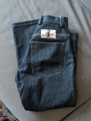Mörkblå jeans från Berlin Denim - Snygga mörkblå jeans från Berlin Denim med klassisk femficksdesign och kontrastsömmar i rött. Jeansen har en cool patch på bakfickan och är tillverkade i robust denim. Perfekta för dig som gillar en stilren och tidlös look.