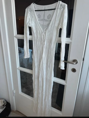 Vit lång strandklänning i virkad stil - Snygg vit strandklänning i virkad stil med lång ärm och djup V-ringning. Klänningen är hellång och har knytning framtill. Perfekt att slänga över bikinin på stranden. Materialet är tunt och luftigt, vilket gör den superbekväm under varma dagar.