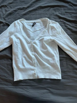 Vit cardigan från Gina Tricot - Snygg vit cardigan från Gina Tricot med v-ringning och knäppning framtill. Långärmad modell i tunt, mjukt material med ribbade muddar och nederkant. Perfekt att slänga över en topp eller klänning.
