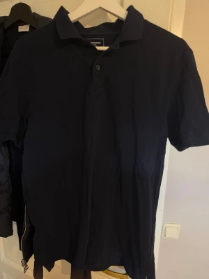 Mörkblå pikétröja med krage - En klassisk mörkblå pikétröja med krage och korta ärmar. Tröjan har knappar framtill och är tillverkad i mjukt bomullsmaterial. Perfekt till jeans eller shorts för en enkel och stilren look.
