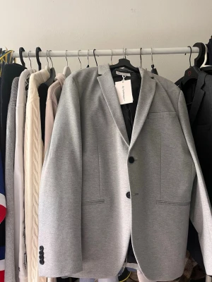 Ljusgrå kavaj från TeeShoppen - Stilren ljusgrå kavaj från By TeeShoppen med två knappar framtill och klassisk krage. Kavajen har släta fickor och svarta knappar vid ärmsluten. Perfekt för en clean och modern look. 