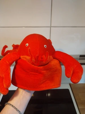 Röd hummerhatt i plysch - Rolig och mjuk huvudbonad i form av en röd hummer med utstickande klor och spröt. Tillverkad i plyschmaterial med svart innerfoder. Perfekt för temafester eller maskerad. Unik accessoar som garanterat drar blickarna till sig.