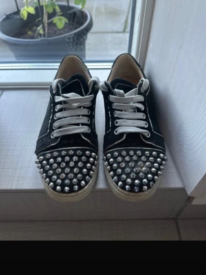 Svarta sneakers med nitar - Svarta sneakers med vita snören och coola silvriga nitar över hela tån och runt hälen. Skorna har en klassisk låg modell och är tillverkade i mockaliknande material med vit sula. Perfekta för dig som vill sticka ut och addera edge till din outfit.