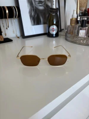 Guldiga retro solglasögon med bruna glas - Snygga solglasögon med tunna guldfärgade metallbågar och bruna, rektangulära linser. Perfekt accessoar för en trendig look med vintagekänsla. Lätta och stilrena, passar till många outfits.