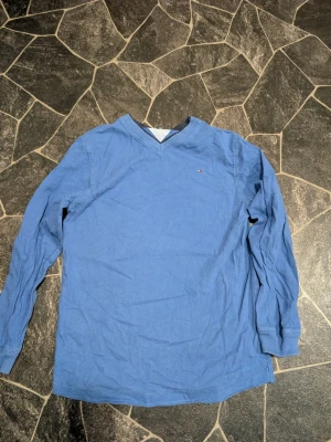 Blå långärmad tröja Tommy Hilfiger stl L 16-18 - Snygg blå långärmad tröja från Tommy Hilfiger med V-ringning och diskret logga på bröstet. Tröjan har ribbade muddar vid ärmslut och hals, perfekt för en avslappnad och stilren look. Stl passar ca 152-160 färgen är lite mörkare än det ser ut på bilderna 