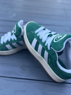 Gröna Adidas Campus sneakers - Gröna Adidas Campus sneakers i mocka med vita klassiska ränder på sidorna och vit sula. Skorna har vit Adidas-logga på hälen och plösen samt vita skosnören. Låg modell med retrovibbar och gummisula. Perfekta för dig som gillar streetstyle och sportig look.