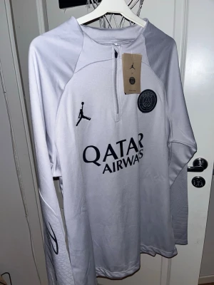 Grå PSG träningströja från Jordan - PRIS KAN DISKUTERAS. Snygg grå långärmad träningströja från PSG x Jordan med halv dragkedja framtill. Tröjan har svarta loggor, PSG-märke på bröstet och Qatar Airways-tryck. Tillverkad i ett lätt och funktionellt material som passar perfekt till träning.