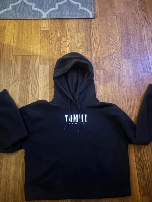 Svart hoodie från Tommy Jeans - Svart hoodie från Tommy Jeans med vit logga på bröstet och klassisk huva. Mjuk insida och ribbade muddar. 