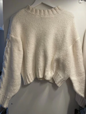 Vit stickad tröja med flätade ärmar - Mysig vit stickad tröja med oversized passform och ribbad krage. Ärmarna har en snygg flätad detalj längs sidorna och tröjan har ribbade muddar vid ärmslut och nederkant. Perfekt för kalla dagar och enkel att matcha med jeans eller kjol.