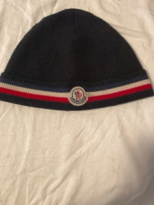 Mörkblå Moncler mössa med ränder - Snygg mörkblå mössa från Moncler med klassiska ribbade ränder i blått, vitt och rött längs kanten. Mössan har Moncler-logga framtill och är stickad i ett mjukt material som håller dig varm under kalla dagar.