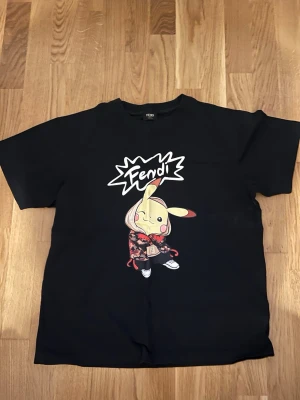 Svart Fendi t-shirt med Pikachu-tryck - Cool svart t-shirt från Fendi med ett stort tryck av Pikachu i streetwear och texten 'Fendi' på bröstet. T-shirten har rund hals och korta ärmar. Perfekt för dig som gillar unika designer och vill sticka ut.