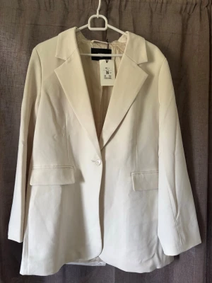 Cremevit blazer - Stilren vit kavaj från bikbok med klassisk krage, enkelknäppning och två fickor framtill. Ärmarna har tre dekorativa knappar vid ärmslutet. Perfekt för en clean och modern look.