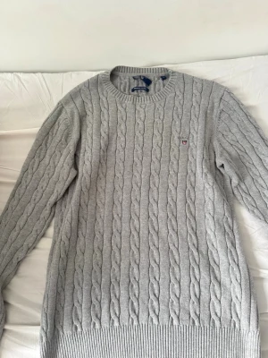 Ljusgrå kabelstickad tröja Gant - Kabelstickad ljusgrå tröja från Gant med rund halsringning och diskret logga broderad på bröstet. Tröjan har långa ärmar och ribbade muddar vid ärmslut och nederkant. Perfekt för lager-på-lager och klassisk stil.