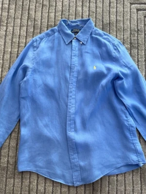 Blå linneskjorta från Polo Ralph Lauren - Snygg blå skjorta i linne från Polo Ralph Lauren med klassisk krage och knappar framtill. Skjortan har lång ärm och den ikoniska broderade loggan i gult på bröstet. Perfekt för en stilren och avslappnad look. Storlek 14-16 år hör av er vid frågor eller funderingar priset går att diskutera!