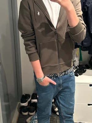 Olivgrön hoodie från Ralph Lauren - Snygg olivgrön hoodie från Ralph Lauren med dragkedja framtill och klassisk huva med dragsko. Ikoniska Polo-loggan broderad på bröstet. Mjukt material och två fickor framtill gör den både stilren och bekväm.