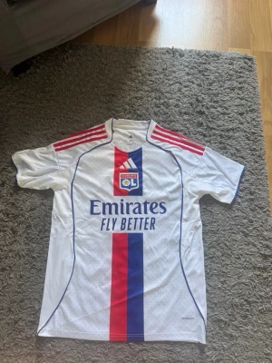 Olympique Lyon fotbollströja Adidas - Säljer en vit Olympique Lyon fotbollströja från Adidas med röda och blå detaljer, klubbmärke på bröstet och sponsortryck 'Emirates Fly Better' framtill. Ryggen har namntryck 'Endrick' och nummer 9. Perfekt för fotbollsfans!