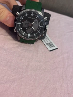 Grön digital- och analogklocka Patterson - Stilren klocka från Patterson med både digital och analog visning. Klockan har en grön syntetrem och svart urtavla med silverfärgade detaljer. Perfekt accessoar för dig som gillar sportig och modern stil.