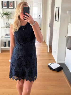 Marinblå spetsklänning med blommönster - Snygg marinblå kortklänning med blommigt spetsmönster och rund halsringning. Klänningen är ärmlös och har en figurnära passform med vacker volangkant nertill. Perfekt för sommarens tillfällen!